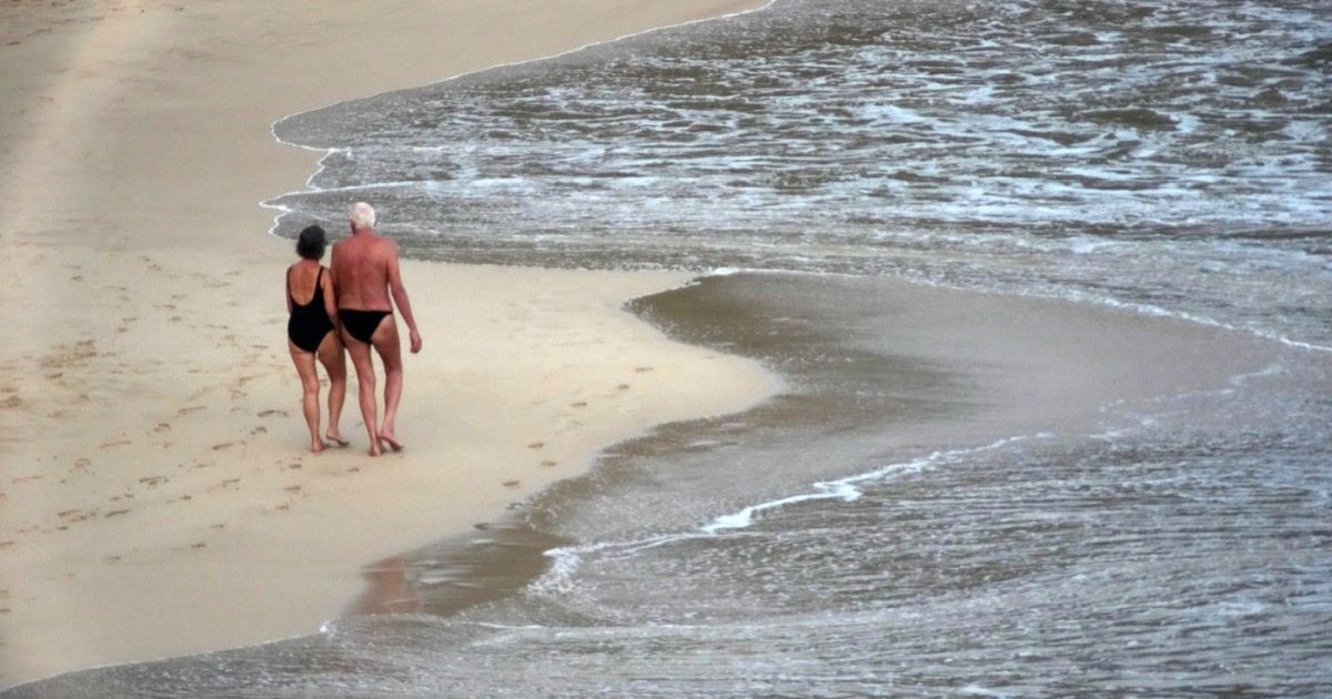 Un cuplu de pensionari în costume de baie se plimbă prin nisipul de la marginea mării, decembrie 2025