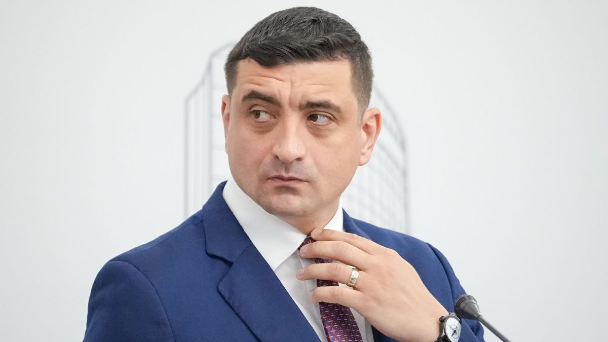 George Simion, îmbrăcat cu un costum albastru, în timp ce își aranjează cravata, pe fundalul unui perete alb