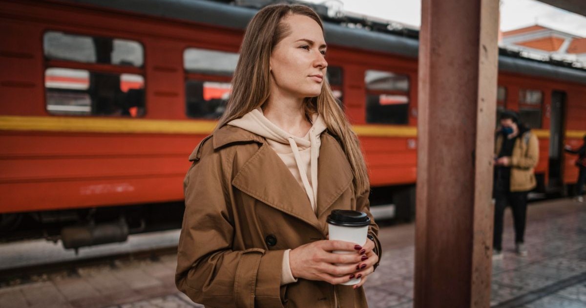 femeie care ține un pahar de cafea într-o gară, cu un tren roșu pe fundal și alți călători