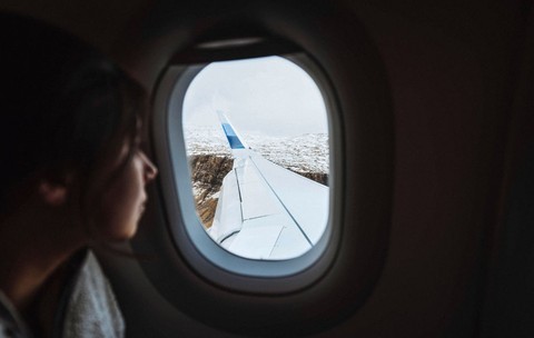 O femeie adultă cu părul închis la culoare privește pe fereastra avionului spre aripa acestuia și spre peisajul de dedesubt, parțial acoperit de zăpadă
