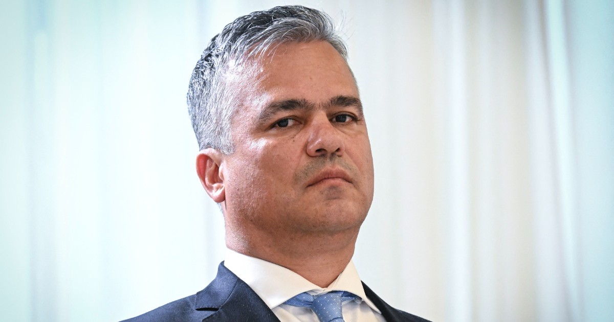 Imaginea prezintă un bărbat îmbrăcat într-un costum închis la culoare și cămașă albă, cu o cravată bleu deschis, surprins într-un cadru oficial. Expresia serioasă și postura sobră sugerează participarea la un eveniment instituțional sau politic.