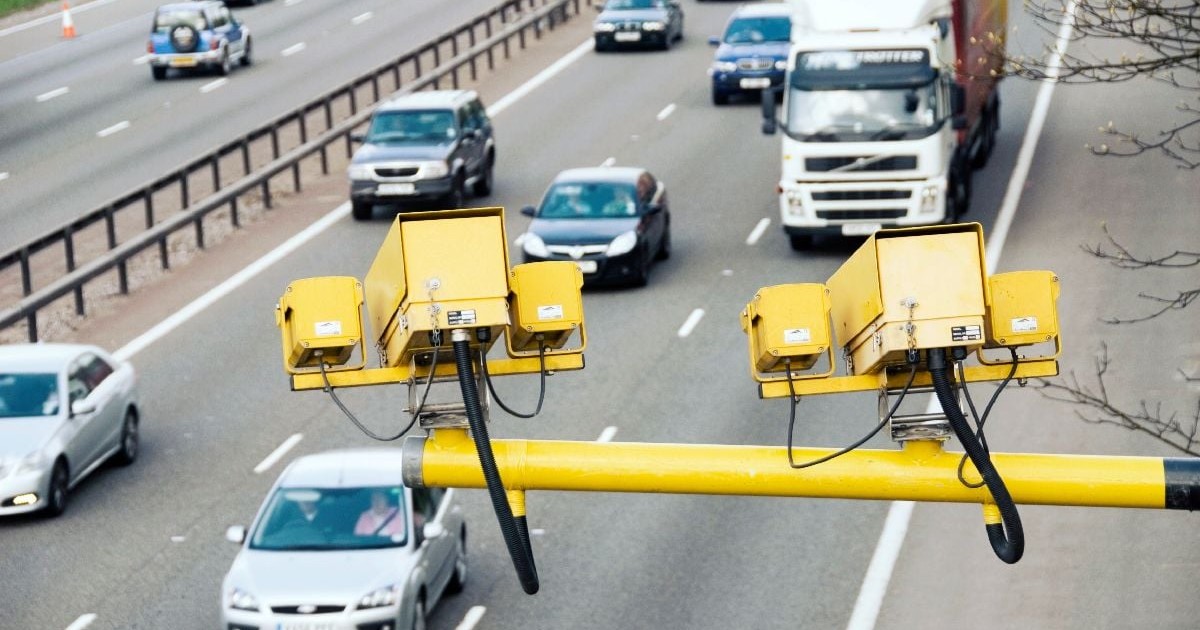 Imaginea prezintă un sistem de camere de monitorizare a traficului, de culoare galbenă, montate deasupra unei autostrăzi aglomerate pe care circulă mai multe autoturisme și un camion.