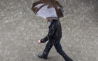 Un bărbat îmbrăcat într-o geacă neagră și pantaloni închiși la culoare, mergând pe un trotuar pavat în timp ce ține o umbrelă maro deasupra capului, pe o ploaie torențială.