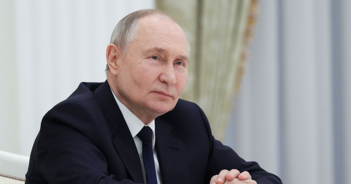 Vladimir Putin la birou, îmbrăcat într-un costum negru cu o cămașa albă și cravata neagră, cu mâinile împreunate așezate pe birou