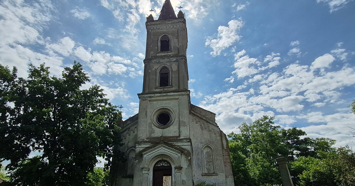 Imagine cu biserica germană ”Sfântul Gheorghe” din satul Malcoci, înconjurată de verdeată iar deasupra ei se vede cerul albastru senin.