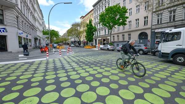 O stradă urbană mărginită de clădiri și copaci, unde carosabilul este pictat cu un model din cercuri galbene de diferite dimensiuni, pe care se deplasează un biciclist.   