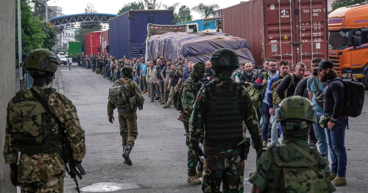 Militari cu spatele supraveghează un șir de bărbați aliniați în dreaptă, lângă mai multe camioane