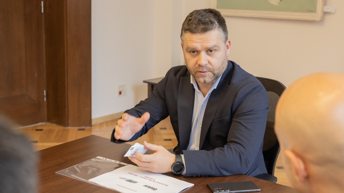 Ciprian Ciucu în costum discută la o masă de birou cu alte persoane, gesticulând, are în față documente și un telefon mobil