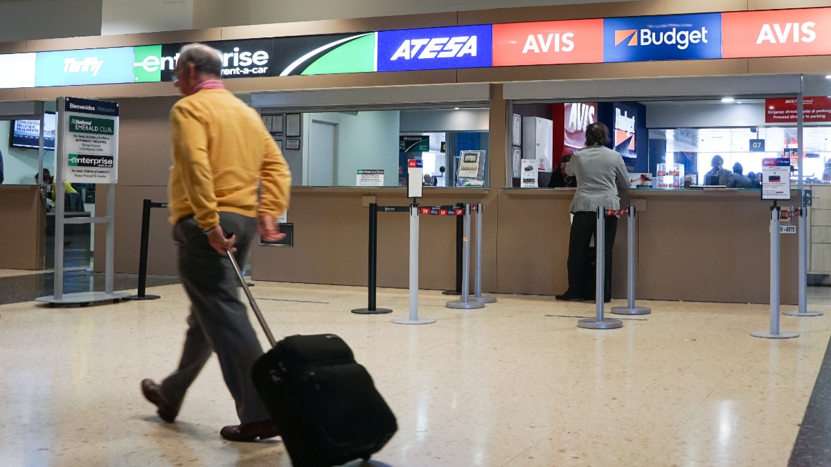 Imaginea arată ghiseele mai multor companii de închirieri auto dintr-un aeroport, în timp ce un bărbat își trage trollerul trecând prin fața lor.