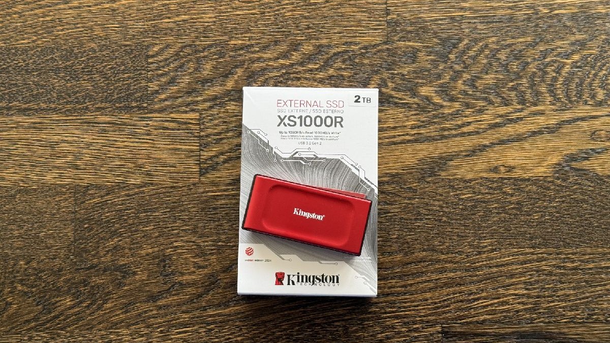 Un SSD extern roșu Kingston XS1000R, așezat pe cutia sa, pe un fundal de lemn.