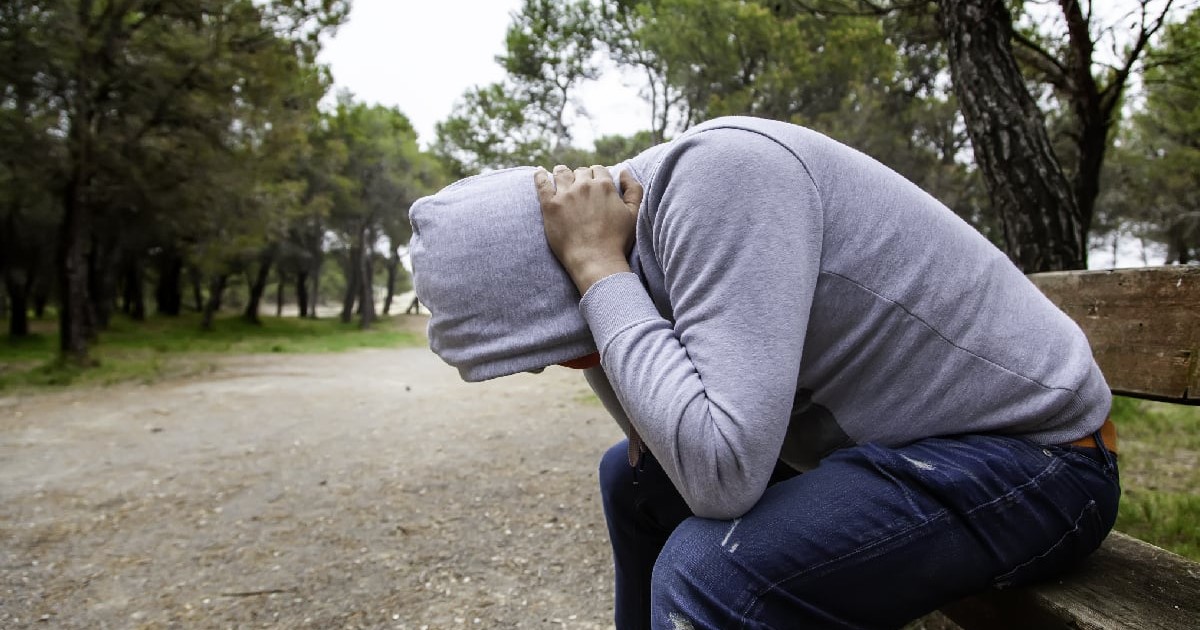 Un tânăr îmbrăcat cu un hanorac se ține cu mâinile de ceafă în timp ce stă pe o bancă într-un parc