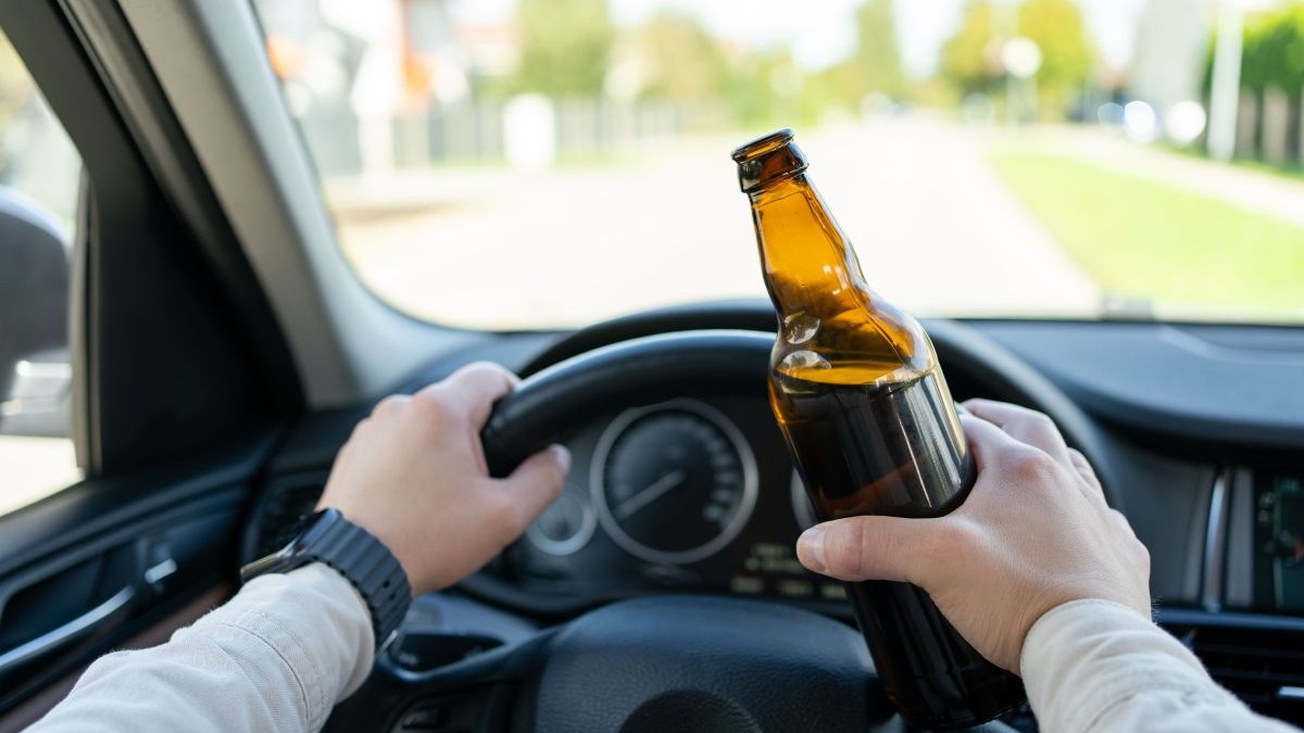Un bărbat conduce o mașină, ține o sticlă de alcool în mână și este cu mâinile pe volanul mașinii