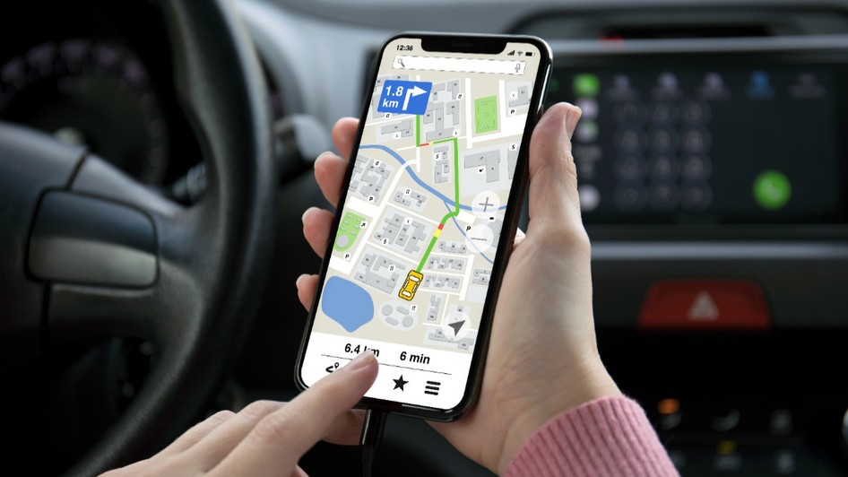 O persoană aflată pe scaunul șoferului al unui autoturism, care ține în mână un smartphone conectat prin cablu și folosește o aplicație de navigare GPS afișată pe ecranul acestuia.