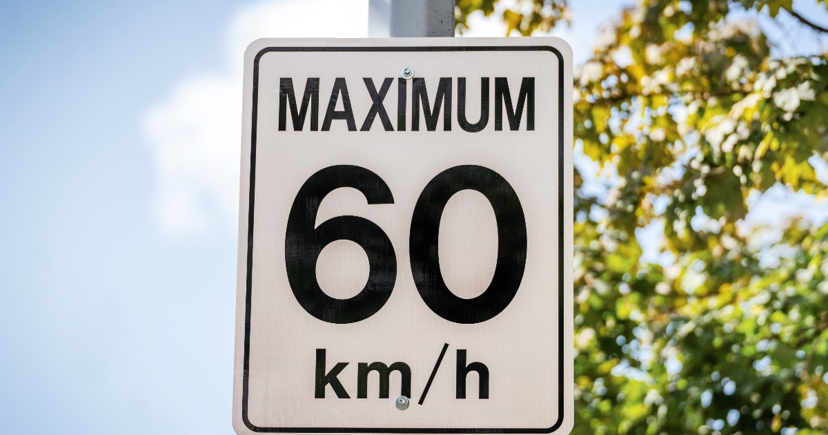 Un indicator rutier de viteză care afișează limita maximă de 60 km/h, montat pe un stâlp metalic, într-o zonă cu vegetație verde în fundal. Semnul are un design simplu, alb cu text negru, specific indicatoarelor de circulație, și sugerează utilizarea sistemului metric pentru măsurarea vitezei. Imaginea este clară, luminoasă și potrivită pentru a ilustra un articol despre sisteme de măsură, trafic rutier sau diferențe între metric și imperial.