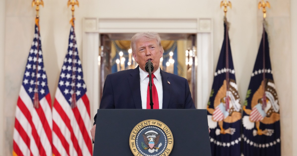 Donald Trump susține un discurs oficial la pupitru, având în spate drapelele Statelor Unite și simbolurile prezidențiale. Imaginea surprinde un moment formal, în care acesta se adresează publicului într-un cadru solemn.