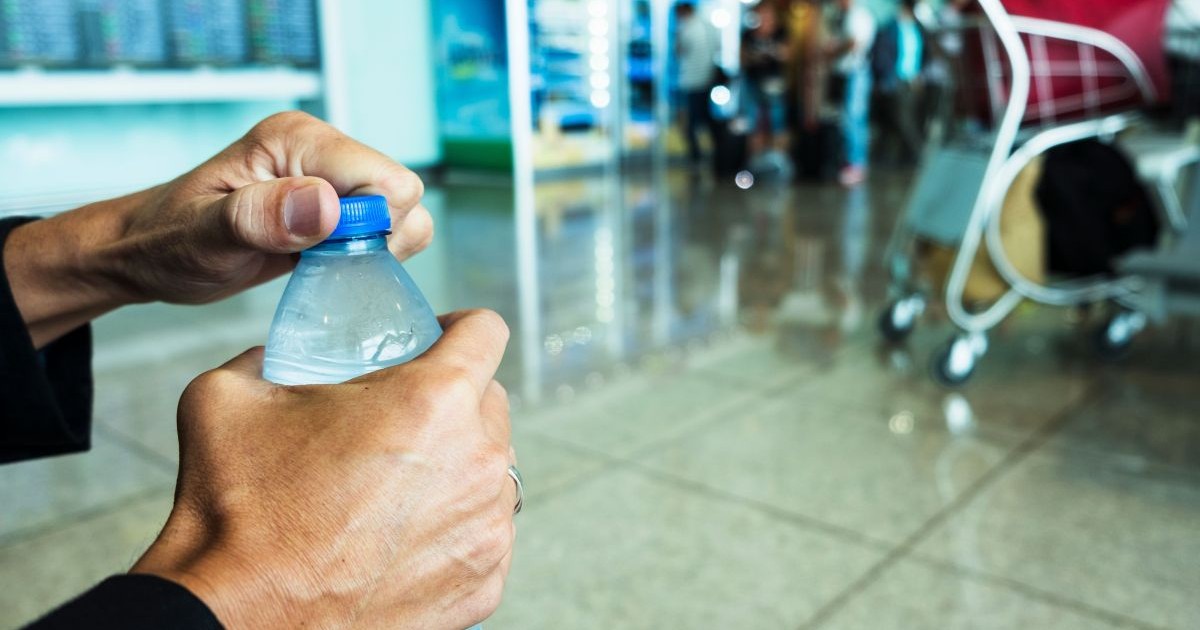 Mâinile unui bărbat care desface o sticlă de apă de 500 de ml, cu dop albastru, iar în fundal se vede o zonă din aeroport și niște begaje