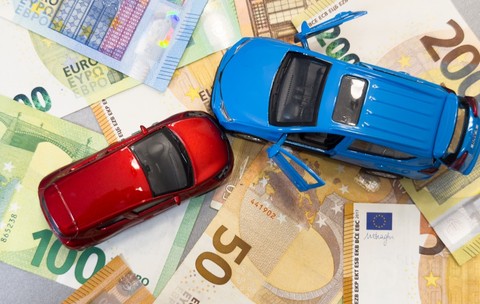 Două mașinuțe de jucărie - una roșie și una albastră - așezate deasupra unui strat de bancnote euro cu cupiuri de 50, 100 și 200 de euro.