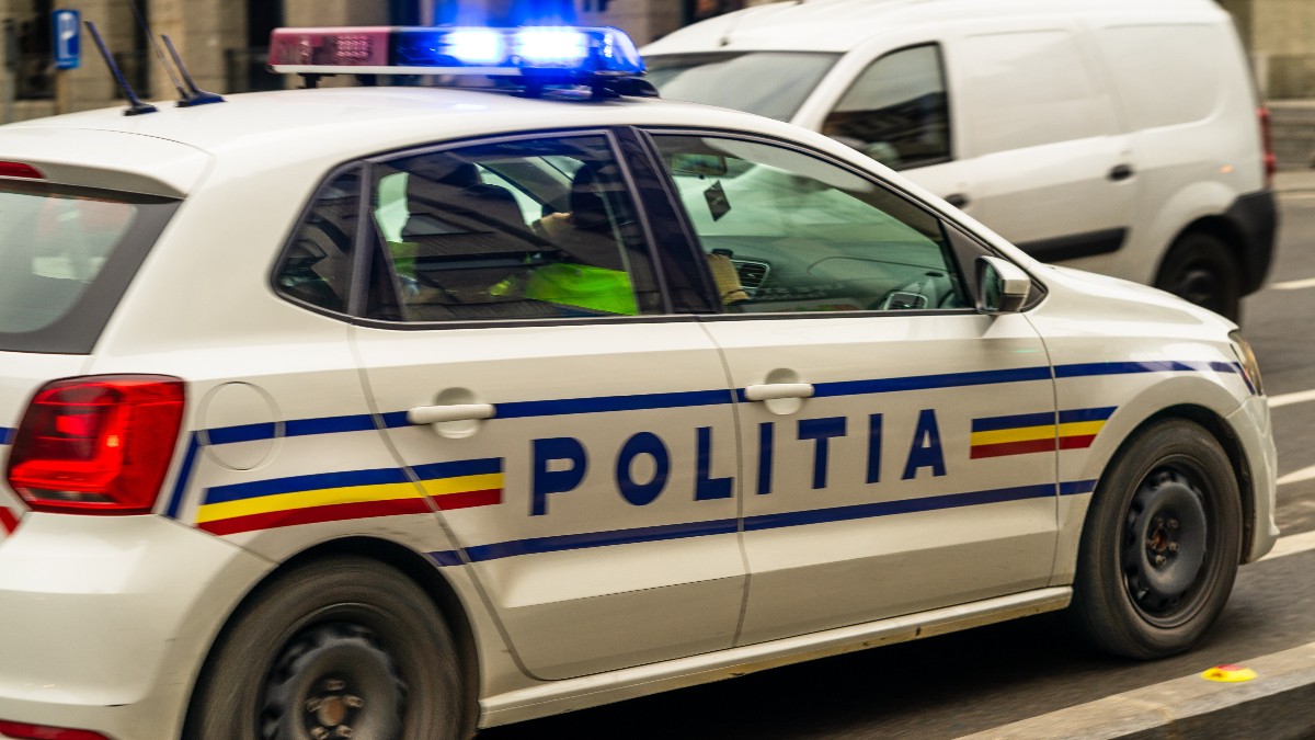 În imagine se vede o autospecială a Poliției Române aflată în trafic, cu girofarurile aprinse. Mașina este inscripționată cu „POLIȚIA” și culorile drapelului României, semnalând o intervenție sau o deplasare oficială.