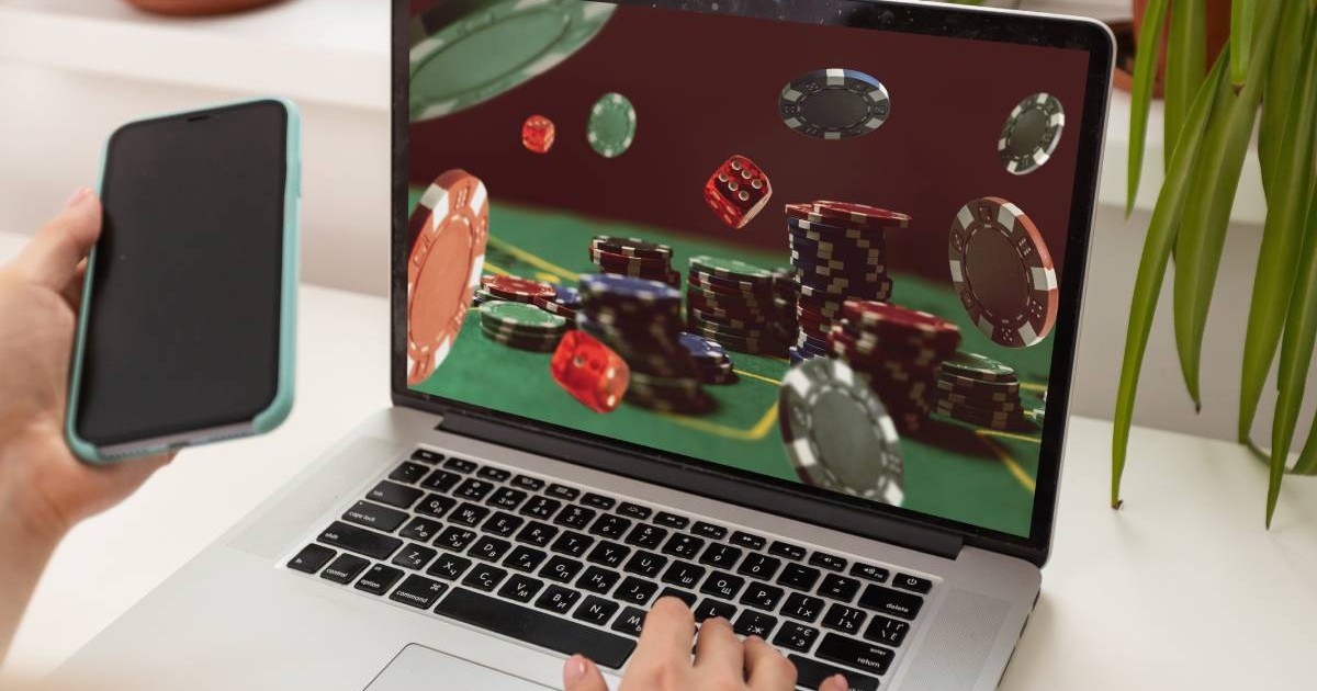 mana unei femei pe tastatura unui laptop deschis pe o pagina de internet cu jocuri de noroc, in cealalta mana are un telefon