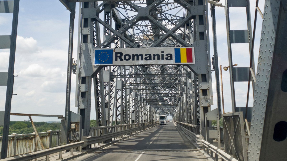 Podul Giurgiu-Ruse, podul Prieteniei, văzut dinspre Giurgiu. Pe șosea se află un TIR alb, iar deasupra ramei metalice ale podului se observă un semn de circulație cu textul „România”, steagul Uniunii Europene și steagul României