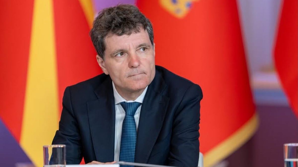 Președintele Nicușor Dan, îmbrăcat la costum, își ține buzele strânse, este la o masă și are în spate un drapel