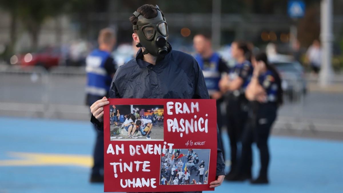 În imagine se vede un protestatar purtând o mască de gaze și ținând o pancartă roșie cu mesajul „ERAM PAȘNICI / AM DEVENIT ȚINTE UMANE”, alături de fotografii de la protestele din 10 august. În fundal se află mai multe persoane, iar scena pare să fie surprinsă în Piața Victoriei, în timpul manifestației comemorative.