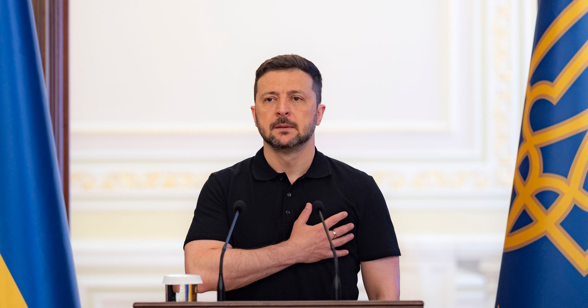Voldimir Zelenski, îmbrăcat într-un tricou polo cu mânecă scurtă, cu mâna stângă la piept, stând la un pupitru de lemn închis la culoare pe care sunt amplasate două microfoane
