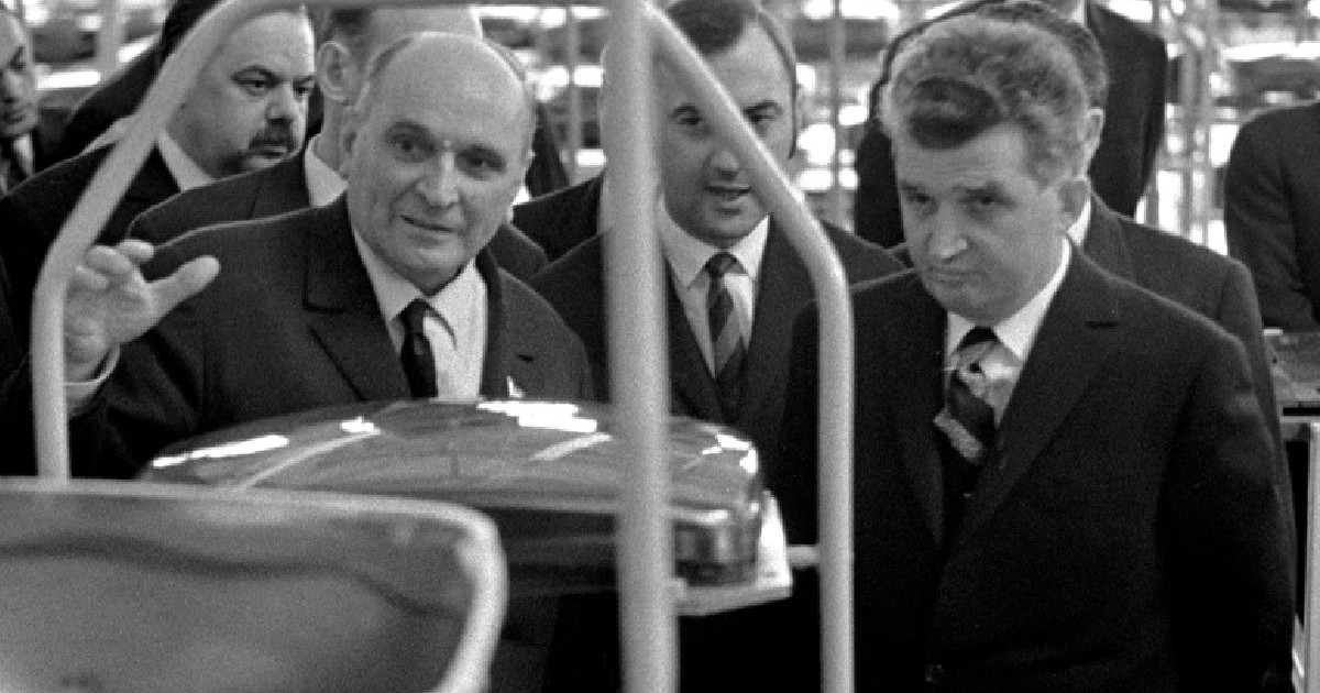 Nicolae Ceaușescu, în dreapta, în costum și cu cravată, ascultă explicațiile unui bărbat în fața unor piese dintr-o fabrică