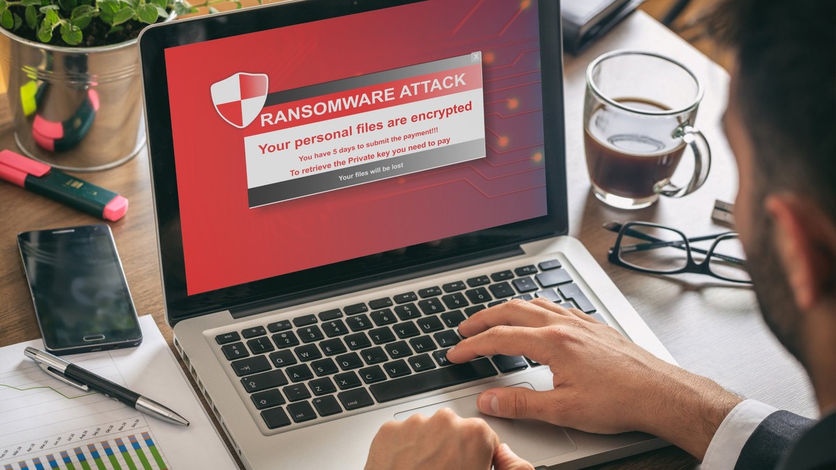 Atacuri Ransomware - Laptop cu imaginea unui atac Ransmoware pe ecran, pus pe un birou, alături de o ceaşcă de cafea, o pereche de ochelari şi obiecte de birotică, agendă, pix, cariocă.