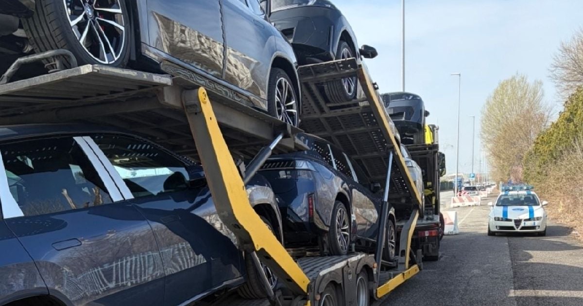 Imaginea arată mai multe mașini așezate pe o platformă staționată pe marginea drumului, cu o mașină de poliție în fundal, pe partea dreaptă a șoselei.