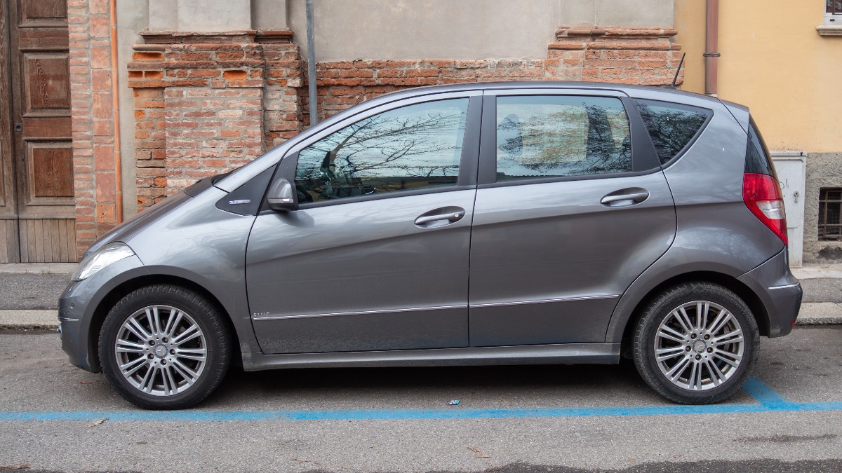 Un automobil Mercedes-Benz Clasa A de culoare gri este parcat pe marginea străzii, pe un loc delimitat cu linii albastre, în fața unei clădiri din cărămidă.