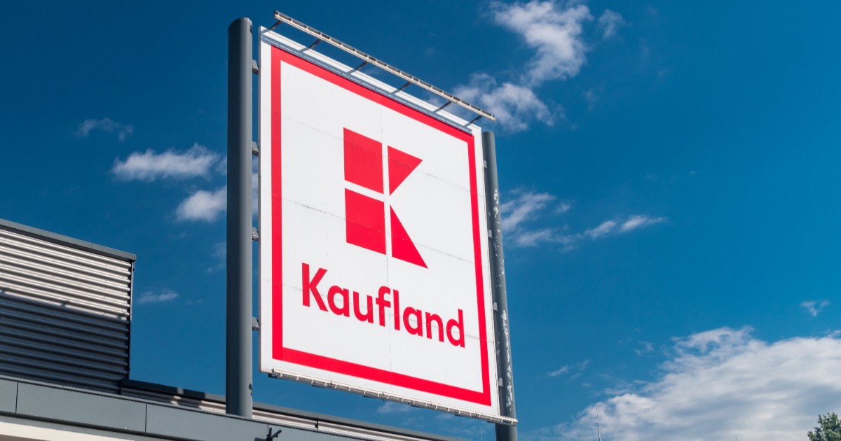 Sigla Kaufland este vizibilă pe fundalul unui cer albastru senin. Panoul este montat pe o structură metalică deasupra unei clădiri.