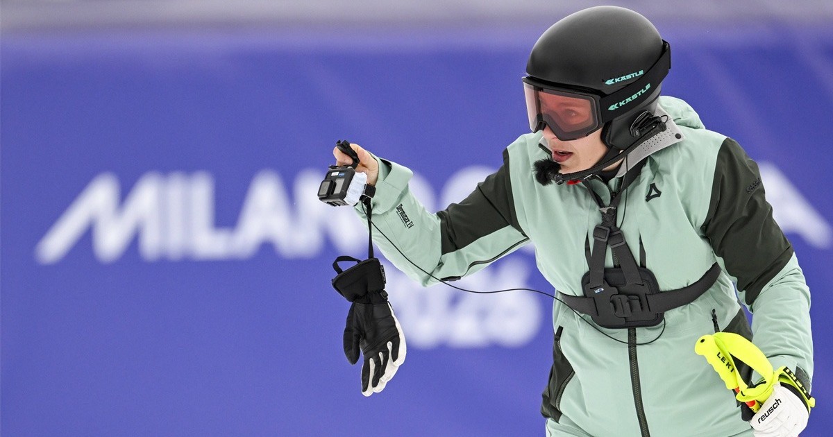O sportiva, schioare, echipata intr-un costum venil, cu o casca de protectie neagra pe cap, cu o manusa atarnand se uita la o mini camera video pe care o tine in mana. Cum rezista sportivii la frig la Olimpiada. Explicatii medic sportiv. Foto Imago