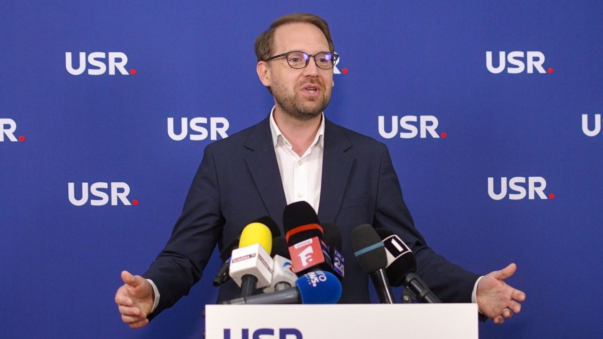 Dominic Fritz îmbrăcat într-un sacou negru și o cămașă albă, vorbind în fața unui pupitru cu sigla USR pe care se află mai multe microfoane, cu mâinile larg deschise