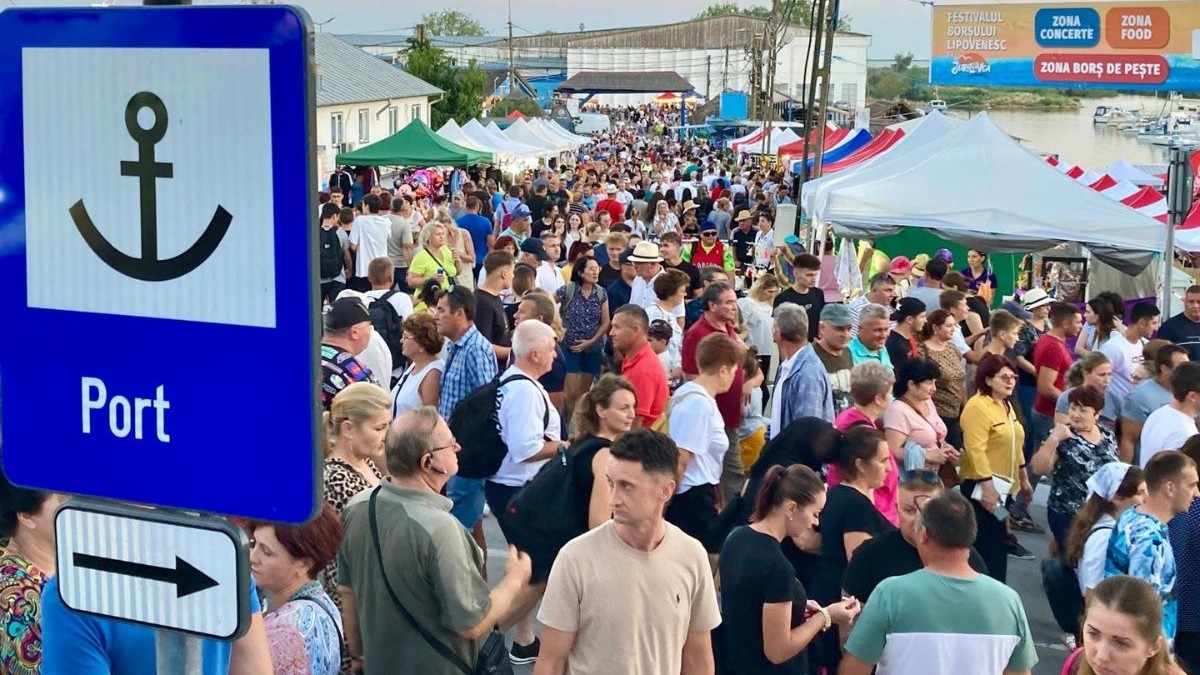 , în partea stângă a prim-planului tronează un indicator mare ce afișează simbolul unei ancore, cuvântul „Port” și o săgeată orientată spre dreapta. Restul cadrului este umplut de o mulțime densă de oameni de diverse vârste care se deplasează pe o alee pietonală largă, participând la un eveniment public foarte aglomerat. De-a lungul traseului, pe ambele părți ale mulțimii, sunt înșiruite numeroase corturi de eveniment. În partea din dreapta sus tronează un panou informativ amplu pe care scrie „FESTIVALUL BORȘULUI LIPOVENESC”, însoțit de direcționări către „ZONA CONCERTE”, „ZONA FOOD” și „ZONA BORȘ DE PEȘTE”. Pe fundal, dincolo de mulțime și de corturi, se conturează clădiri cu acoperișuri înclinate, stâlpi de electricitate și o întindere de apă cu ambarcațiuni ancorate la mal.