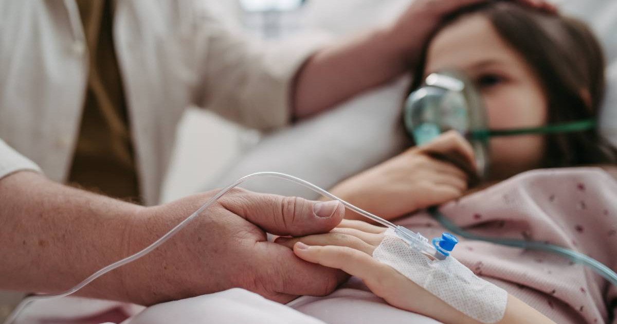 Mana unui barbat in halat alb tine de mana o fetita, aflata pe patul de spital, cu o masca de oxigen pe fata,