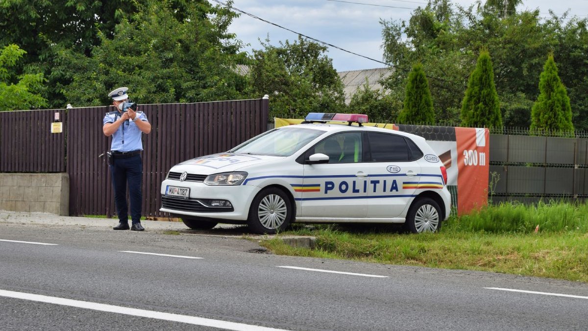 Polițist îmbrăcat în uniformă de vară, cu cămașă albastră și mânecă scurtă, cu un radar în mână, în picioare, lângă mașina de Poliție, parcată lângă un gard de lemn, cu vegetație verde pe fundal, lângă o șosea