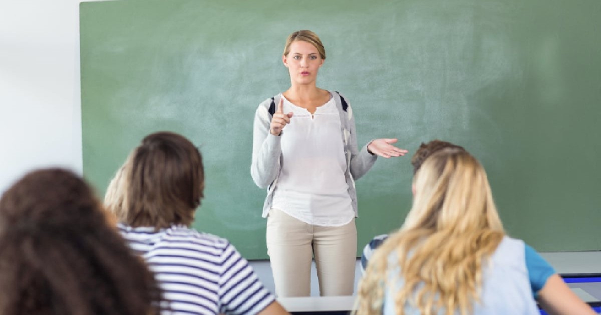 Imaginea prezintă o profesoară stând în fața unei clase de elevi. Ea este îmbrăcată casual, cu o cămașă albă, un cardigan gri deschis și pantaloni bej. Are părul blond, prins, și o expresie facială serioasă, cu o mână întinsă și un deget ridicat, ca și cum ar explica sau accentua un punct important. Cu cealaltă mână, face un gest deschis, sugerând o discuție. În fundal se află o tablă de culoare verde, iar în prim-plan, se văd din spate capetele elevilor, așezați la pupitre. Unii dintre ei au capetele întoarse, ascultând-o pe profesoară. Atmosfera generală a imaginii este de studiu și de predare-învățare, specifică unui mediu școlar.