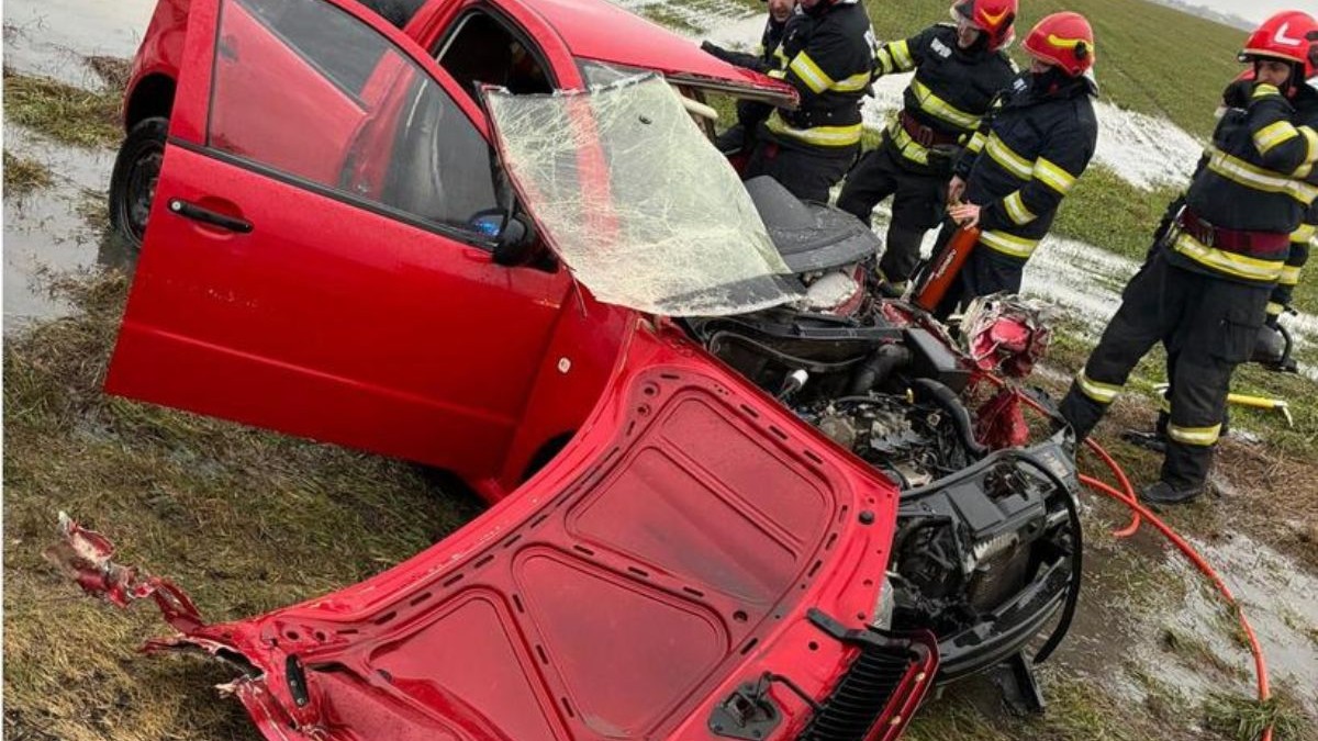 masina rosie cu capota sarita, parbrizul spart si grav avariata frontal, dupa un accident, inconjurata de pompieri in uniforme negre si casti rosii pe cap