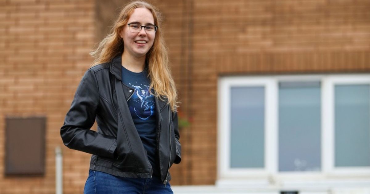 Charlotte Sprason, studenta care și-a cumpărat casă la 24 de ani, în fața casei sale, îmbrăcată cu blugi de culoare albastră, o bluză închisă la culoare și o geacă de piele neagră