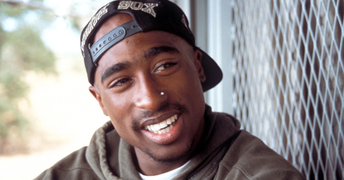 Imaginea prezintă un portret de aproape al celebrului rapper Tupac Shakur, zâmbind și purtând o șapcă orientată în spate și un hanorac verde.