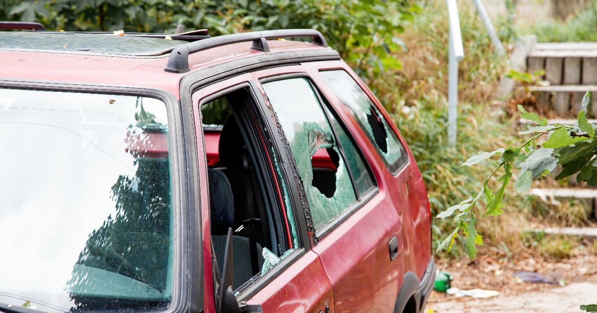 Imaginea prezintă un autoturism de culoare roșie care a suferit avarii semnificative, având mai multe geamuri laterale sparte. Mașina este parcată într-o zonă cu vegetație, iar urmele distrugerilor sunt vizibile atât la nivelul geamurilor, cât și al caroseriei