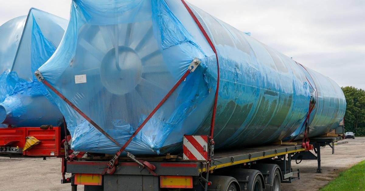 Remorcă platformă care transportă un rezervor industrial supradimensionat din oțel inoxidabil, învelit în plastic albastru pentru protecție, fixat cu curele de siguranță