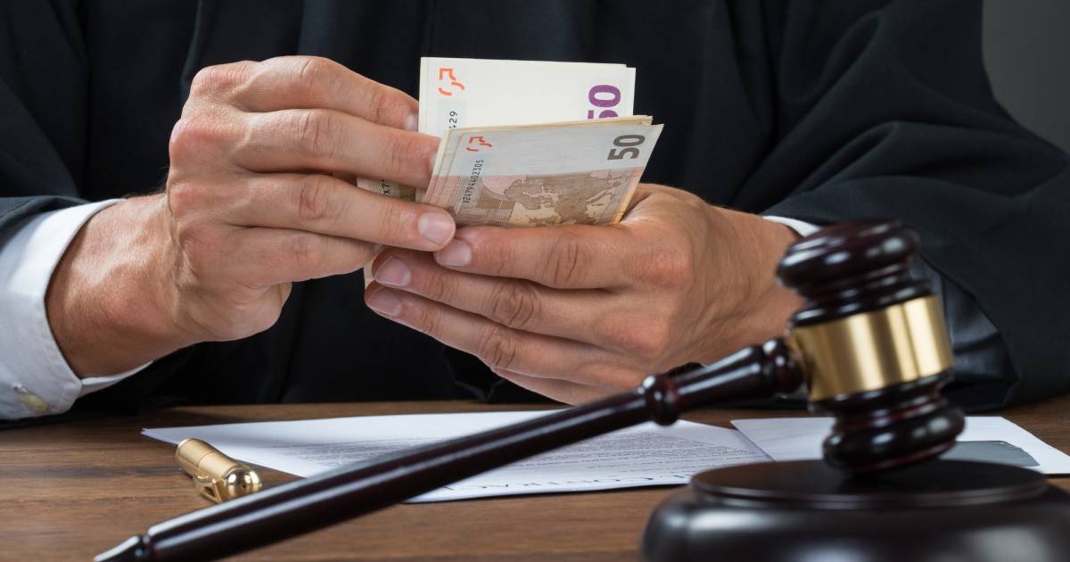 Mainile unui judecator care numara mai multe bancnote de 50 de euro, in fata, pe masa, foi albe si un stilou, in prim plan ciocanelul judecatorului,