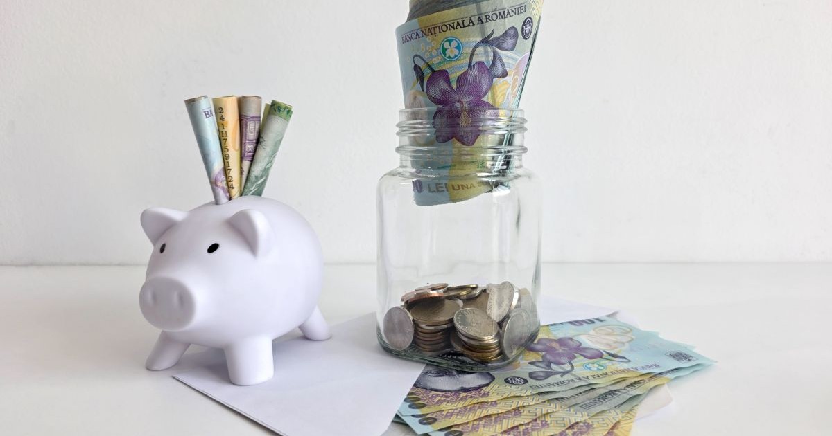 O pușculiță albă în formă de porc, plină de bani românești, sub ea este un plic alb umplut cu bancnote de 100 de lei, iar peste el este un borcan transparent din sticlă plin cu monede, iar deasupra hârtii de 100 de lei