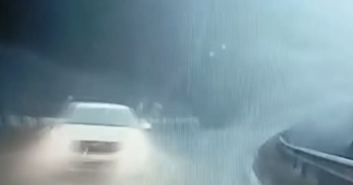 Imaginea surprinde o scenă pe un drum umed și încețoșat. O mașină albă se deplasează cu farurile aprinse dinspre stânga spre centru. Drumul face o curbă spre dreapta, cu un parapet vizibil pe partea dreaptă.