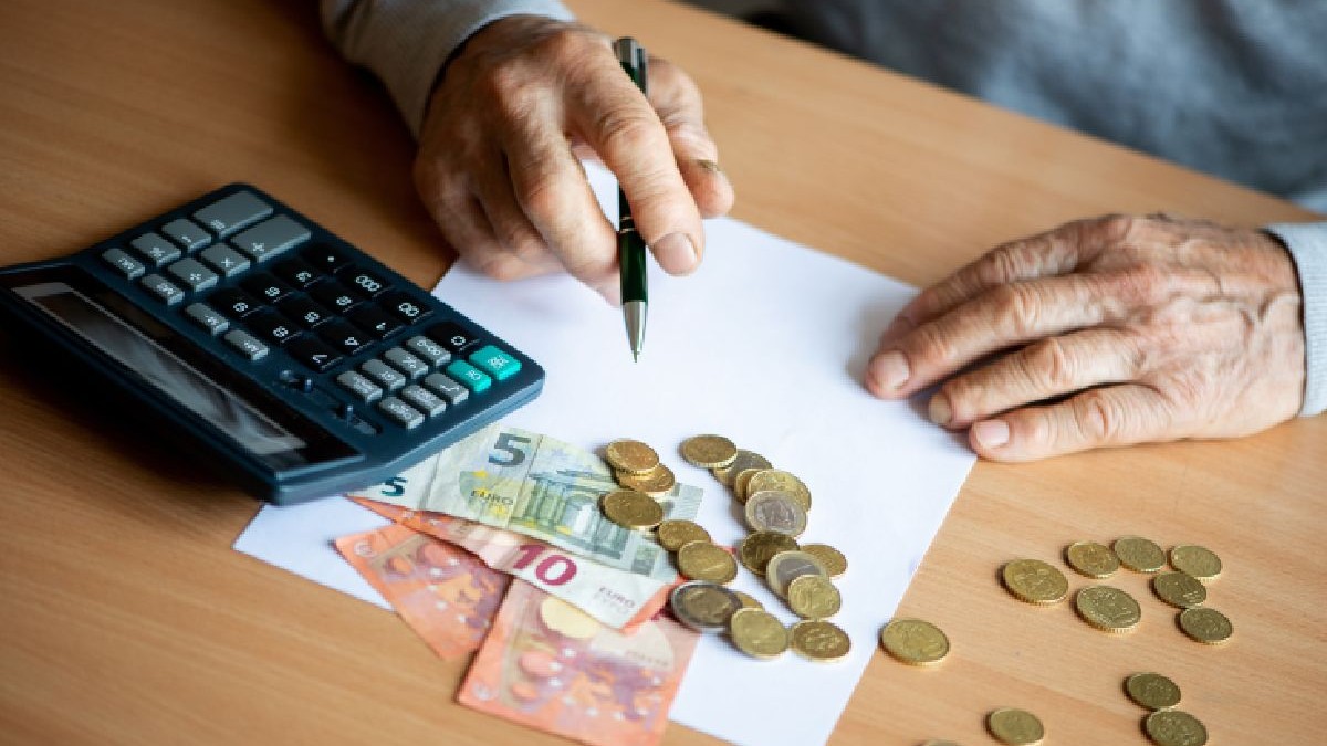 O fotografie arată o persoană în vârstă scriind pe o foaie albă de hârtie cu un stilou. Persoana este așezată la o masă de lemn. Un calculator mare și o grămadă de bancnote euro sunt așezate în stânga foii. Monede sunt împrăștiate în jurul foii, inclusiv monede euro de diferite valori.