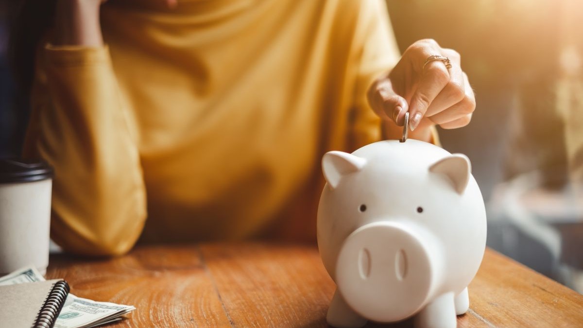 O femeie pune bani într-o pușculiță de tip porc alb, aflată pe o masă. Foto cu caracter ilustrativ: Shutterstock