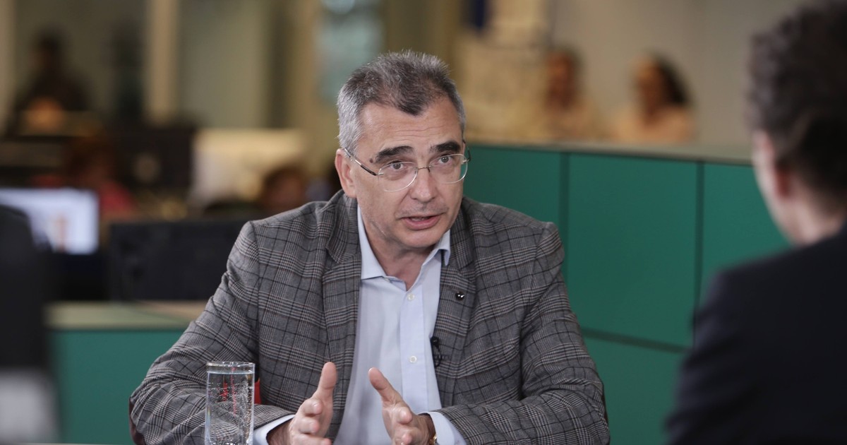 Petrișor Peiu, îmbrăcat într-un sacou crem cu model de carouri și o cămașă albă, stând la masă cu coatele pe casă și palmele împreunate. alături de mâna sa stângă se află un pahar cu apă pe jumătate plin