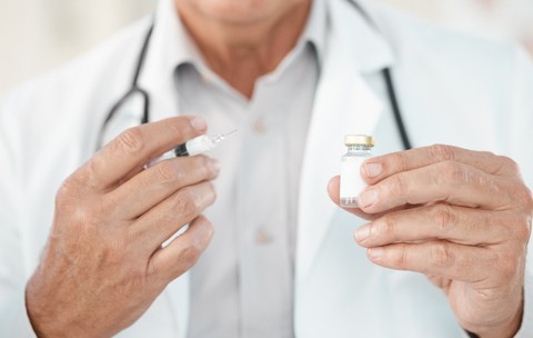 Un doctor îmbrăcat în halat alb, purtând un stetoscop în jurul gâtului, care ține o seringă cu ac și un flacon mic de sticlă